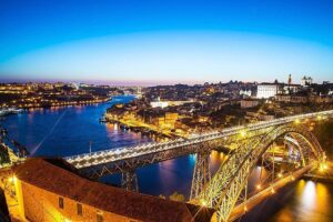 Photo prise du côté des caves à vin donnant sur le quai de Vila Nova de Gaia, offrant une vue sur les bateaux traditionnels Rabelo, le pont ancestral Dom Luís I de style Eiffel, et le quartier historique de Porto sur la rive du Douro, capturée en fin d'après midi.