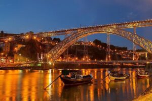 Photo prise du côté des restaurants et caves à vin sur le quai de Vila Nova de Gaia, offrant une vue sur les bateaux traditionnels Rabelo, le pont ancestral Dom Luís I de style Eiffel, et le quartier historique de Porto sur la rive du Douro, capturée de nuit.