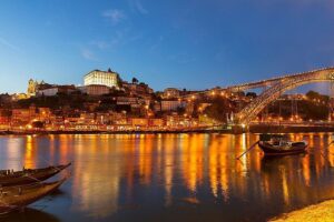 Photo prise du côté des restaurants et caves à vin sur le quai de Vila Nova de Gaia, offrant une vue sur les bateaux traditionnels Rabelo, le pont ancestral Dom Luís I de style Eiffel, et le quartier historique de Porto sur la rive du Douro, capturée de nuit.
