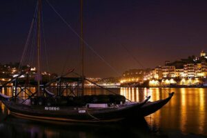 Photo nocturne du bateau traditionnel Rabelo, utilisé autrefois pour le transport des vins de la région du Douro, sur la rive de Vila Nova de Gaia, avec l'éclairage nocturne mettant en valeur le paysage pittoresque le long du Douro.
